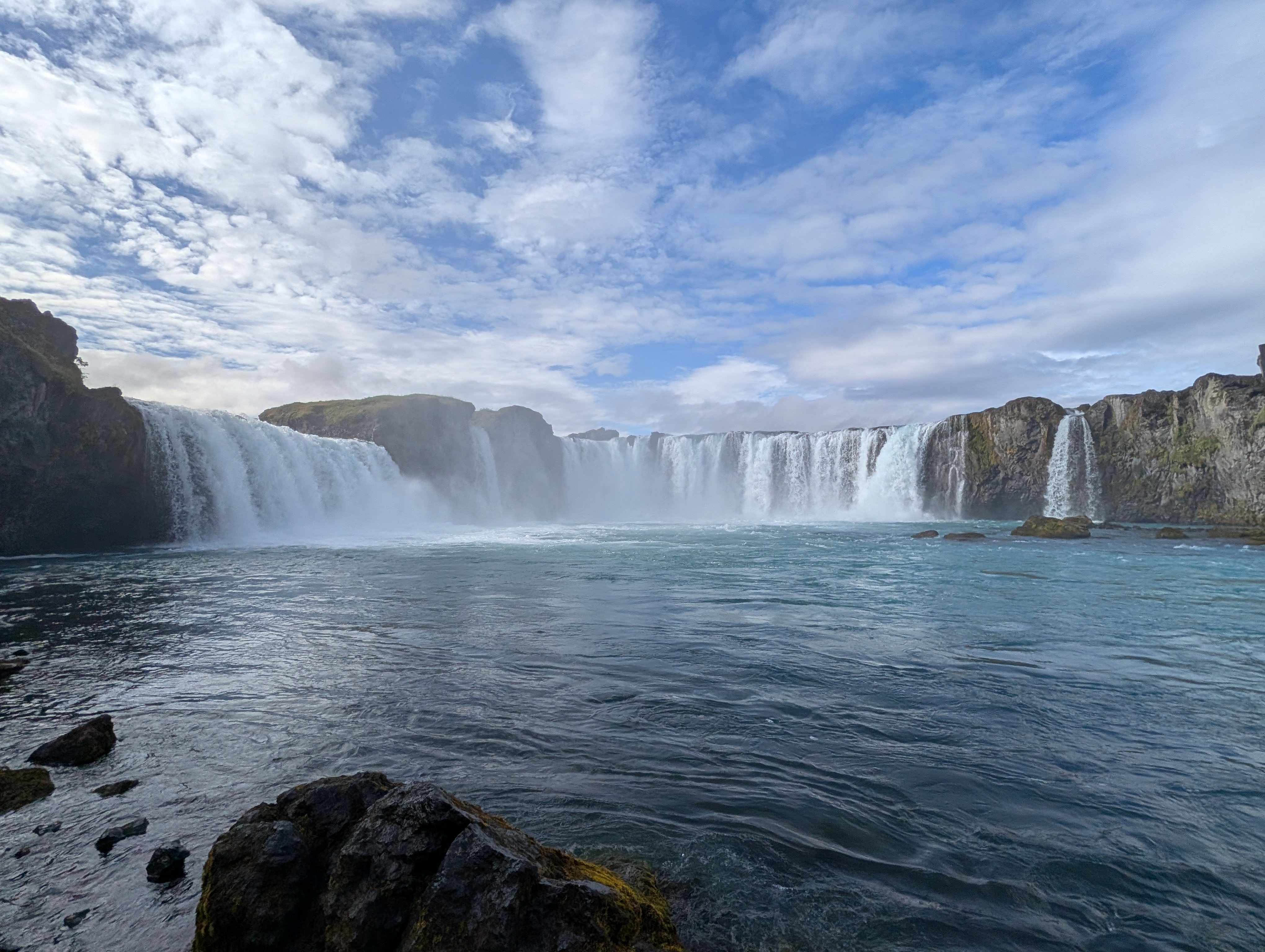 Goðafoss waterfall flowing over a broad rocky ledge beneath a blue, cloud-filled sky